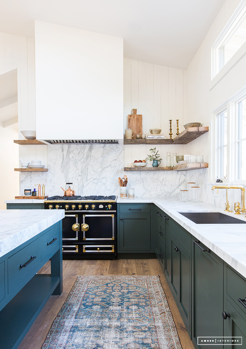 Dark green cabinets with marble backsplash, French Oven, and wood open shelving via Amber Interior Design