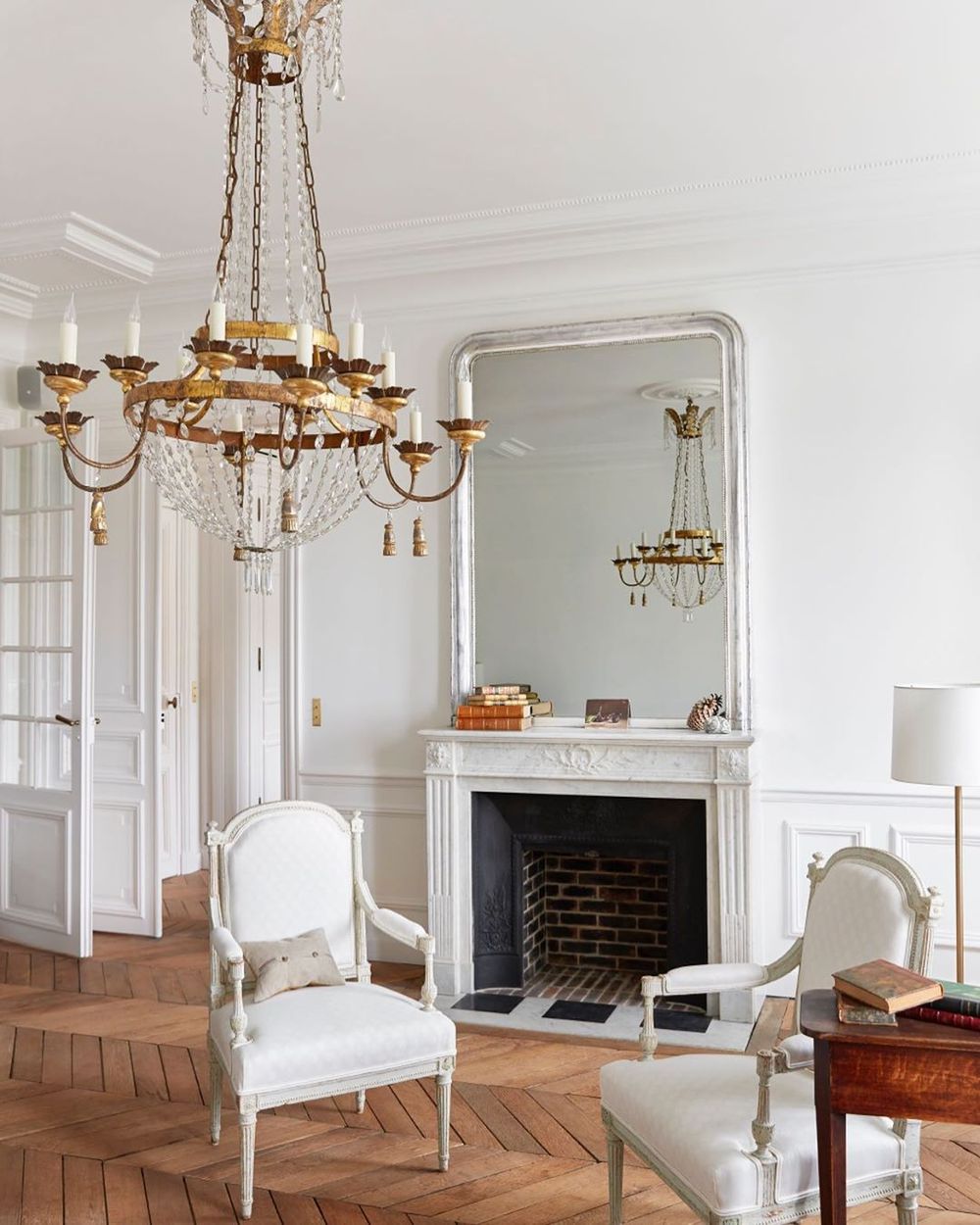 White neoclassical Parisian fireplace with two white French sitting chairs, parquet flooring and crystal chandelier via @abkasha